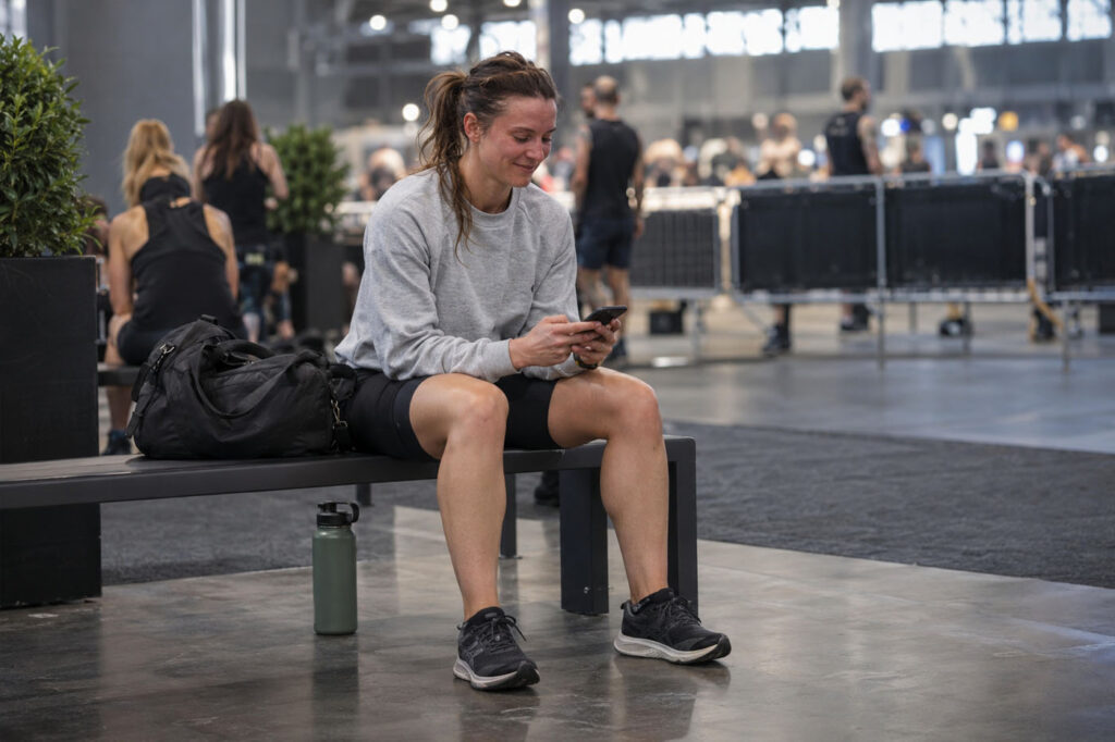 Female athlete sitting in a race venue rest area, checking her phone after completing a HYROX competition.