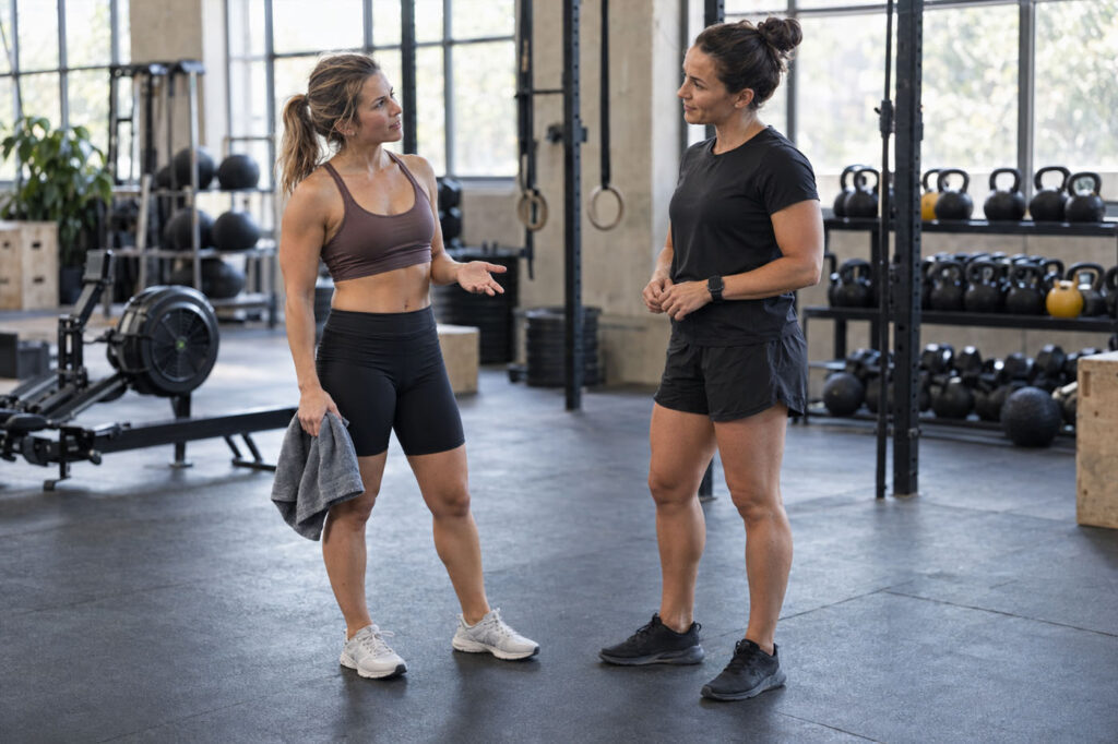 Female athlete speaking with a coach beside a gym training area before joining a HYROX class.